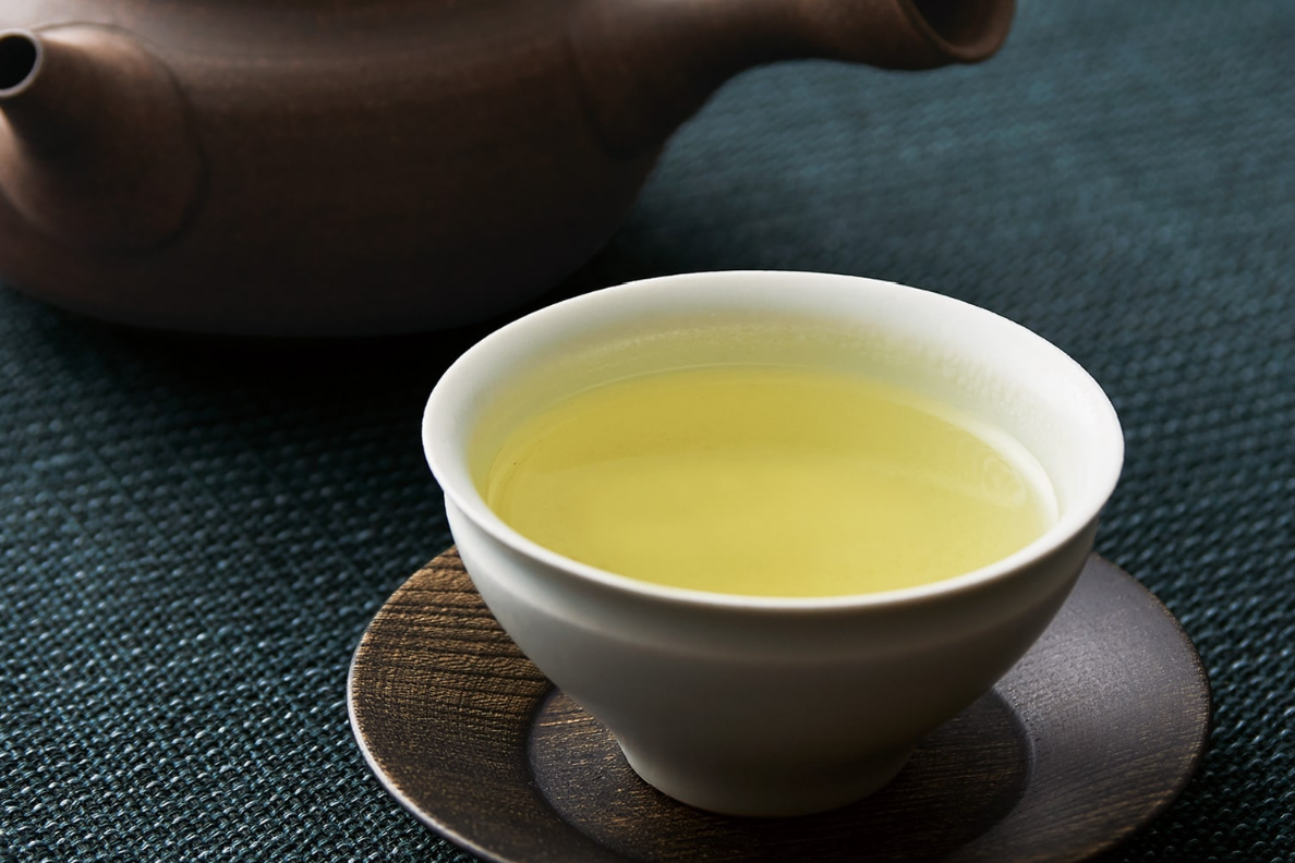White cup of green tea with a brown teapot on a textured surface. UZUKI